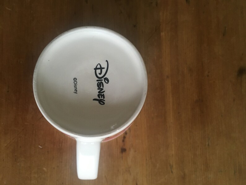 Peut inclure: Mug en c&eacute;ramique blanche avec une anse, portant le logo Disney et le symbole du copyright. Le mug est pos&eacute; sur une surface en bois. L'int&eacute;rieur du mug est blanc.