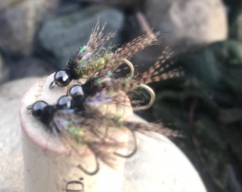 May include: Close-up of several fishing flies with black beads and iridescent feathers, resting on a cork. The flies are designed for fly fishing. The background is blurred, showing water and stones.