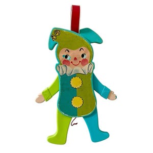 May include: A vintage wooden toy in the shape of a jester. The jester is painted in shades of green and blue, with yellow accents. It has a red ribbon for hanging. The jester's face is painted with a smiling expression.