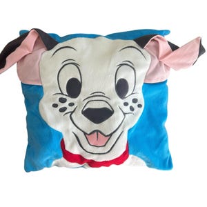 May include: A square plush cushion with a cartoon dog's face. The dog has a white face with black spots, black eyes, and a pink tongue. The ears are pink and black, and the cushion has a blue background and a red collar.