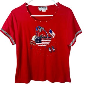 Puede incluir: Camiseta roja de manga corta con un diseño patriótico. La camiseta presenta una cesta con un diseño de bandera estadounidense, estrellas y un lazo. Las mangas tienen rayas blancas y azul marino.