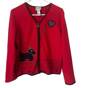 May include: A red zip-up cardigan with a black dog and heart design. The sweater has a black trim and a zipper with a heart-shaped pull. The dog is on a leash that leads to a heart. The brand tag reads "Napa Valley".