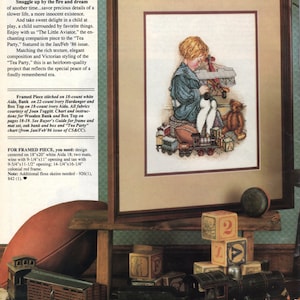 Könnte beinhalten: Ein gerahmtes Kreuzstichstück mit dem Titel "The Little Aviator" zeigt ein Kind umgeben von Spielzeug, darunter ein Teddybär, eine Eisenbahn und Blöcke. Das Kunstwerk befindet sich in einem braunen Rahmen mit einem beigen Passepartout, was einen Vintage-Look erzeugt.