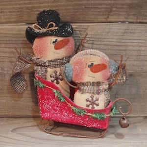 Puede incluir: Dos muñecos de nieve de tela hechos a mano en un trineo rojo con una campana. Los muñecos de nieve llevan sombreros y bufandas y tienen caras pintadas. El trineo está decorado con ramitas verdes.