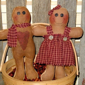May include: Two handmade gingerbread-style dolls with button eyes and stitched smiles. One doll wears a scarf and has a heart on its chest. The other doll wears a plaid dress and a bow. They are in a wooden basket filled with berries.