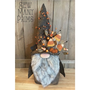 May include: A black and brown gnome with a long, pointed hat decorated with fall foliage, candy corn, and berries. The gnome has a long, gray beard and a black nose.