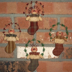 May include: Three handmade Christmas stocking ornaments with a gingerbread person design. Each stocking is decorated with a green wreath and red berries. The stockings are hanging on a brick wall.