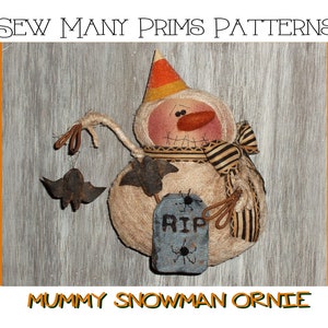 May include: A handmade mummy snowman ornament with a candy corn hat, a striped scarf, and a tombstone that says "RIP". The snowman is holding a stick with two bats hanging from it.