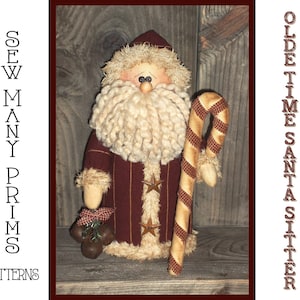 Op de afbeelding: Een handgemaakte, vintage-stijl kerstmanpop met een witte baard en een rood-gouden gestreepte snoepstok. De pop zit op een houten oppervlak en draagt een rood-gouden gestreepte jas. De tekst "OLDE TIME SANTA SITTER" is zichtbaar aan de rechterkant van de afbeelding.
