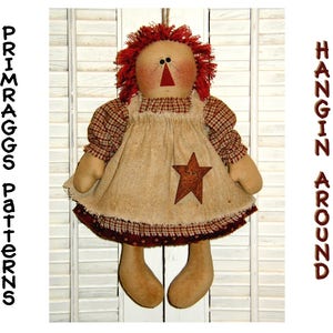 May include: A handmade rag doll with red hair, a brown gingham dress, and a brown star on her apron. The doll is hanging from a white wooden slat wall. The text "PRIMRAGGS Patterns" is on the left side of the image and "HANGIN AROUND" is on the right side of the image.