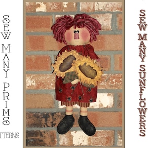 Op de afbeelding: Een roodharige lappenpop met een rode jurk en een geruit overhemd houdt twee zonnebloemen vast voor een bakstenen muur. De tekst "SEW MANY PRIMS PATTERNS" staat aan de linkerkant van de afbeelding en "SEW MANY SUNFLOWERS" aan de rechterkant van de afbeelding.