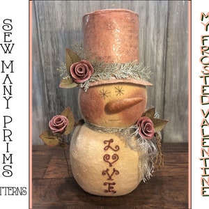 May include: A pink and white snowman with a pink top hat and a brown nose. The snowman is holding a pink rose and has the word "LOVE" embroidered on its chest.
