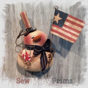 Puede incluir: Un muñeco de nieve patriótico con un sombrero y una bufanda rojo, blanco y azul, sosteniendo una pequeña bandera estadounidense. El muñeco está hecho de tela y tiene un aspecto rústico y vintage. "Sew Many Prims" está escrito en la parte inferior de la imagen.