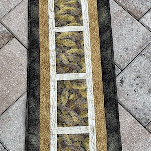 Può includere: Un runner da tavolo rettangolare lungo con un motivo geometrico nero, oro e bianco. Il runner presenta cinque pannelli rettangolari con una stampa di aghi di pino marroni e verdi.