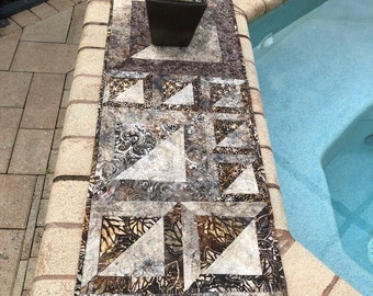 May include: A patchwork table runner with a geometric design in brown, beige, and gold tones. The runner is rectangular and features repeating triangular and square patterns. A plant in a dark pot sits on the runner, which is placed on a stone ledge.