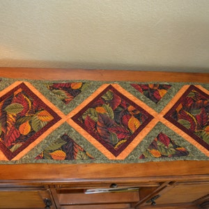 May include: A table runner with a geometric pattern of orange, green, and brown leaves. The runner is made of fabric and has a quilted texture.