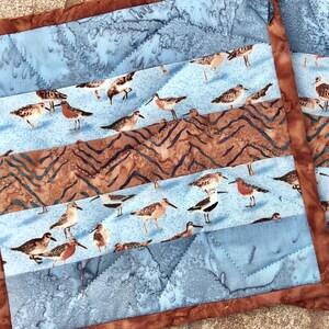 May include: A blue and brown quilted fabric with a pattern of birds. The fabric is made up of several strips of different colors and patterns, and the birds are arranged in a random pattern.