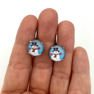 May include: A pair of silver dangle earrings with a round glass charm featuring a snowman wearing a black top hat, red scarf, and red mittens. The snowman is standing in a snowy scene with a blue background.