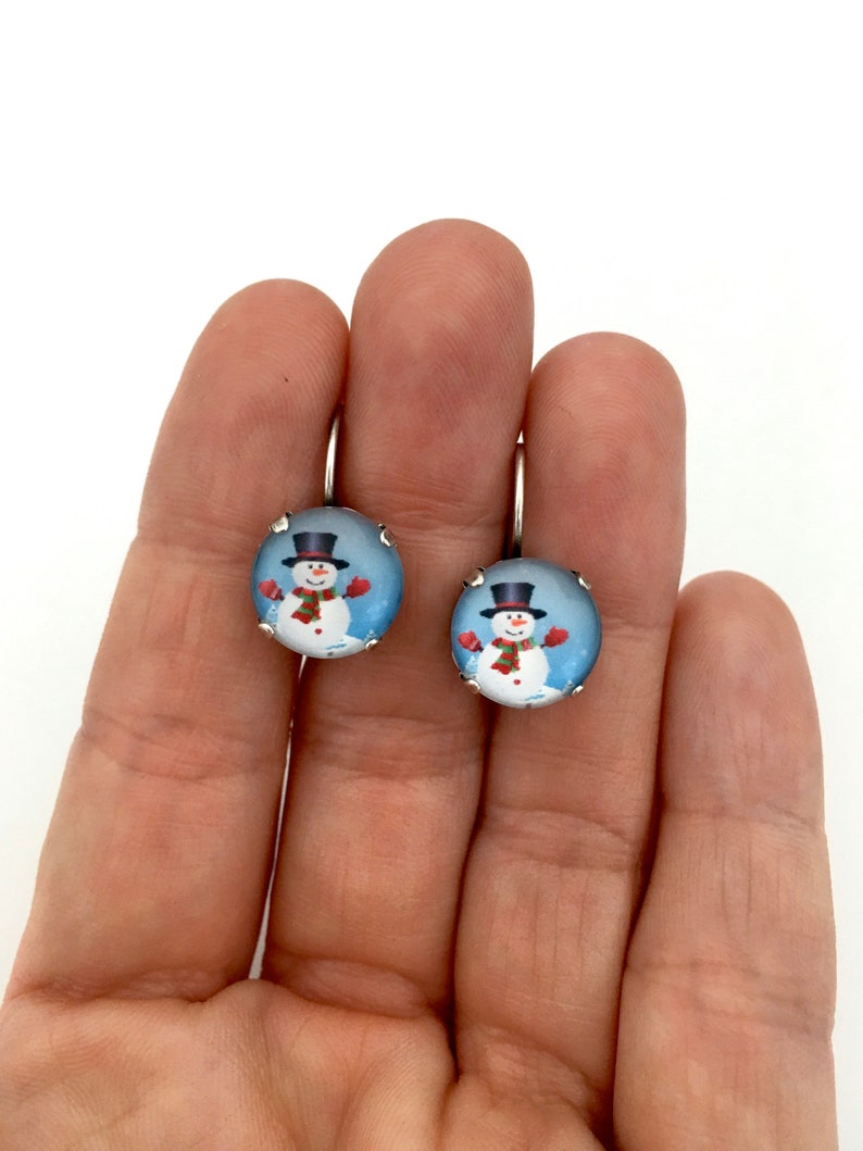 May include: A pair of silver dangle earrings with a round glass charm featuring a snowman wearing a black top hat, red scarf, and red mittens. The snowman is standing in a snowy scene with a blue background.