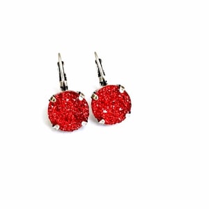 May include: A pair of leverback earrings featuring round, sparkling red druzy stones. The earrings have silver-colored metal settings and leverback closures, set against a white background. These earrings are a bold statement piece.