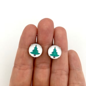 May include: A pair of silver earrings with a round, white background and a green Christmas tree design.