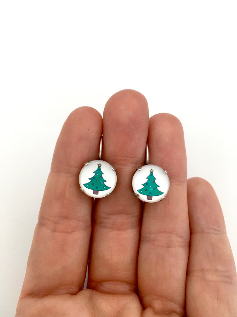 May include: A pair of silver earrings with a round, white background and a green Christmas tree design.