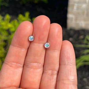 May include: Two small, round stud earrings with iridescent, crystal-like centers. The earrings are held in an open hand against a blurred background of green foliage and sunlight. The earrings appear to be made of metal.