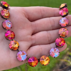Puede incluir: Un collar con cristales redondos e iridiscentes en tonos rosa, naranja y amarillo. Los cristales están engarzados en un marco de metal oscuro y ensartados para formar un collar. El collar se muestra sobre un fondo verde borroso.