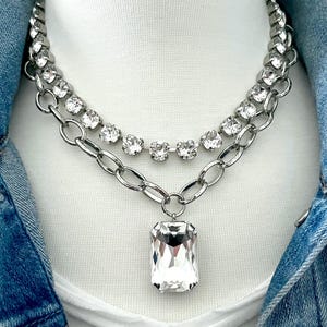 K&ouml;nnte beinhalten: Eine silberne Kettenkette mit einem gro&szlig;en, klaren, rechteckigen Kristallanh&auml;nger. Die Kette ist mit einer kleineren Kette aus klaren Kristallen geschichtet.