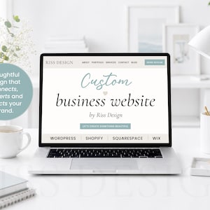 May include: A laptop displays a website design with the text "Custom business website by Riss Design." The screen is surrounded by a white desk with a coffee cup, notepad, and a gold pen. A lamp and flowers are in the background.