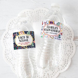 以下が含まれることがあります： ラベルが付いた透明なウォーターボトルが2本。1つのラベルには、学校をテーマにしたカラフルなイラストとともに「BACK TO SCHOOL」と書かれています。もう1つのラベルには「CHARLES ELEMENTARY FALL OPEN HOUSE」と書かれています。どちらのラベルも濃い青色の背景です。