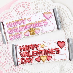 May include: Two chocolate bar wrappers with a Valentine's Day theme. The wrappers are white with pink and gold hearts, glitter, and the text "Happy Valentine's Day". One wrapper has a gold crown and the other has a red heart.