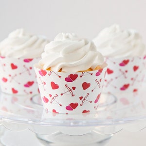 Puede incluir: Tres cupcakes con glaseado blanco sobre una fuente de cristal transparente. Los envoltorios de los cupcakes son blancos con un patrón de corazones rojos, corazones rosas y flechas de Cupido. Los cupcakes son un postre.