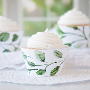 May include: Three cupcakes with white frosting sit in decorative paper cups. The cups are white with a green watercolor eucalyptus leaf design. The cupcakes are on a white surface, with a soft-focus background.