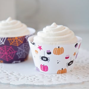 May include: Two cupcakes with white frosting in decorative Halloween-themed wrappers. One wrapper is purple with orange and pink spiderwebs. The other is white with orange pumpkins, spiders, and stars.