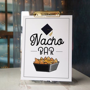 Puede incluir: Un cartel blanco con texto negro que dice "Nacho Bar" con un icono de gorro de graduación sobre el texto. El cartel está en un soporte de acrílico transparente y tiene un clip dorado en la parte superior.