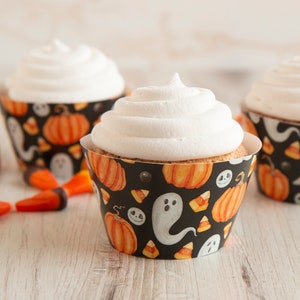Puede incluir: Tres cupcakes con glaseado blanco en envoltorios de papel con temática de Halloween. Los envoltorios son negros con calabazas naranjas, fantasmas blancos y diseños de caramelos de maíz. Caramelos de maíz esparcidos alrededor.