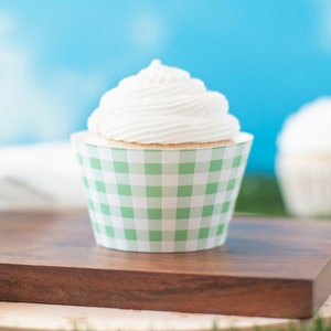 Könnte beinhalten: Ein einzelner Cupcake mit weißem Frosting in einer grün-weißen Gingham-Cupcake-Form. Der Cupcake steht auf einem Holzbrett.