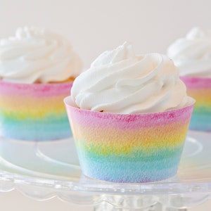 Op de afbeelding: Drie cupcakes met witte frosting en regenboogkleurige cupcake-vormpjes. De vormpjes hebben een aquareleffect met tinten roze, geel, blauw en groen.