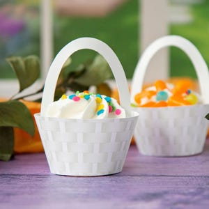 Puede incluir: Dos soportes para cupcakes de papel blanco con forma de cestas con asas. Una cesta está llena de glaseado blanco y chispas de colores, la otra con caramelos naranja y amarillos.