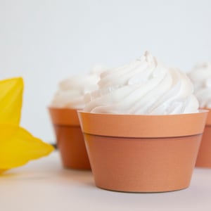 May include: Three white frosted cupcakes in brown flower pot cupcake liners. The cupcakes are arranged in a row on a white surface. A yellow flower is in the foreground.