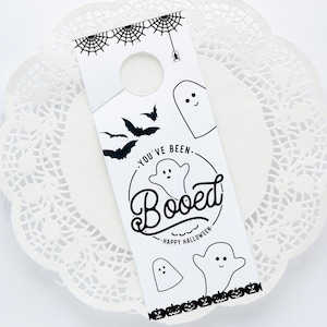 May include: A white door hanger with a black spiderweb design, bats, and ghosts. The text reads "You've Been Booed! Happy Halloween".