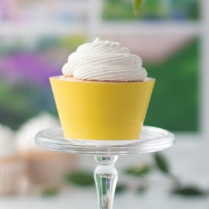 Puede incluir: Un solo cupcake con glaseado blanco en un forro de cupcake amarillo sobre un soporte para pastel de vidrio transparente.