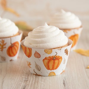 May include: Three cupcakes with white frosting and fall-themed cupcake liners. The liners feature orange pumpkins and brown leaves on a white background.
