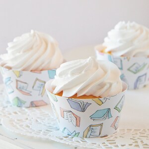 May include: Three white frosted cupcakes with a book-themed design on the cupcake liners. The liners have a white background with colorful illustrations of open books.