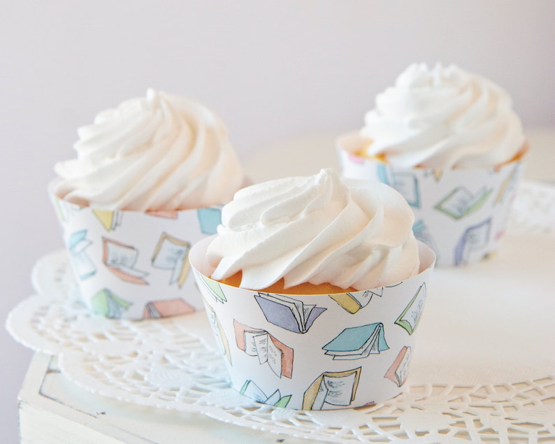 May include: Three white frosted cupcakes with a book-themed design on the cupcake liners. The liners have a white background with colorful illustrations of open books.
