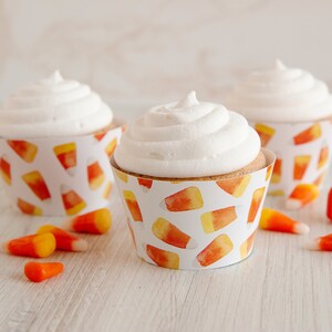 May include: Three cupcakes with white frosting sit in candy corn-patterned wrappers. Candy corn and a small pumpkin-shaped candy are scattered around the cupcakes on a white wooden surface. The cupcakes are a festive treat.