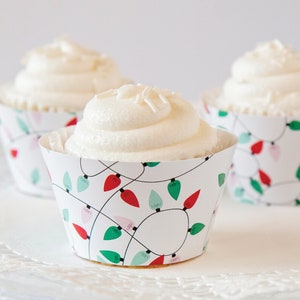 May include: Three white cupcakes with white frosting and sprinkles, decorated with festive Christmas lights cupcake liners. The liners are white with red, green, and pink lights.