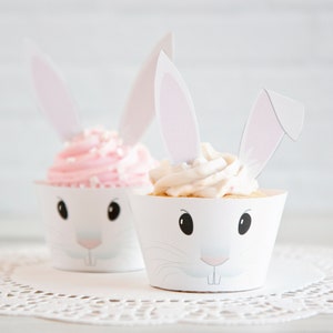May include: Two cupcakes decorated with bunny ears. The cupcakes are white with pink and white frosting. The cupcake wrappers are white with a bunny face design.
