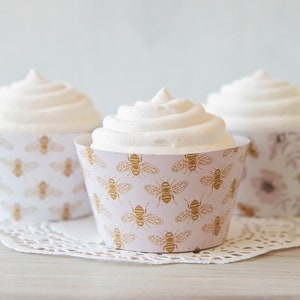 May include: Three cupcakes with white frosting and decorative cupcake liners. The liners have a pink background with a gold bee pattern.